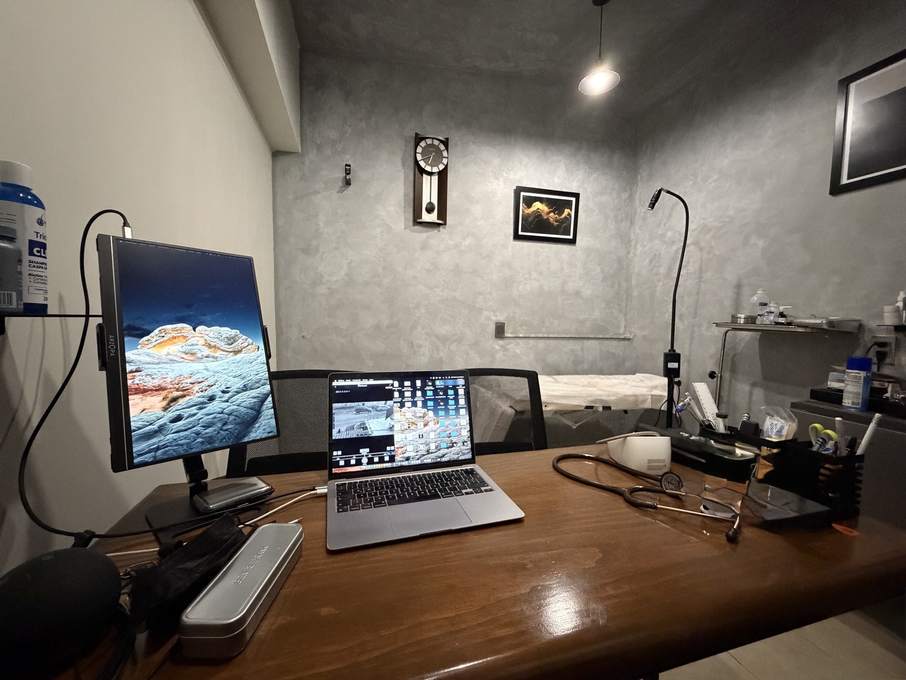 Escritorio del consultorio con monitor, estetoscopio y área de exploración al fondo